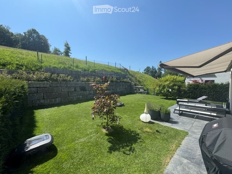 Well-maintained garden with a lawn, a small tree, potted plants, outdoor furniture including a bench and a table, and a covered patio area.
