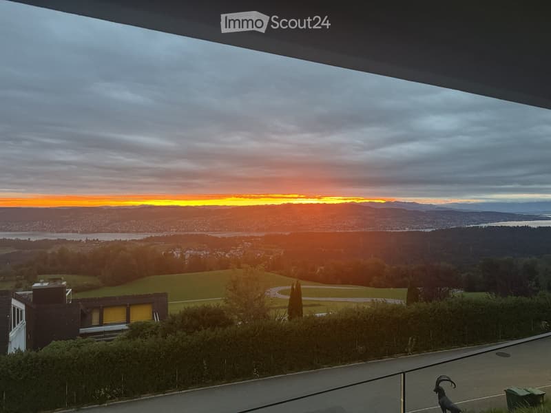 Panoramic view from a balcony or terrace showcasing a cityscape, lake, and mountains during sunset.