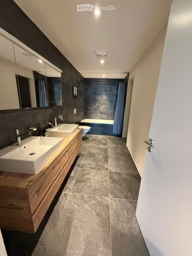 Modern bathroom with double sinks, large mirror, bathtub, and toilet. Grey tiles and wooden vanity.