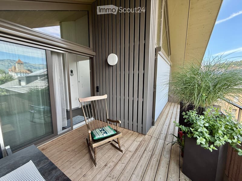Balcony with wooden decking, rocking chair with cushion, potted plants, and scenic city view.