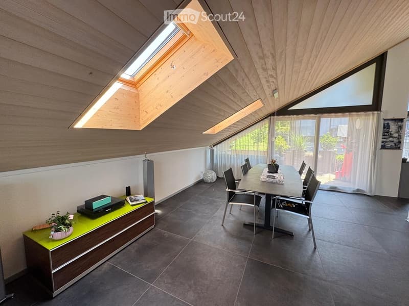 Modern dining area with a sleek dining table, chairs, a sideboard with a plant, and a laptop on top. The room features a skylight and large windows, with a view of the garden.