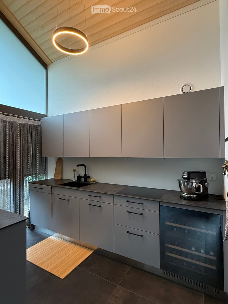 Modern kitchen with grey cabinets, a sink, a mixer, and a wine cooler. Features a large window with a curtain and a circular light fixture on the ceiling.