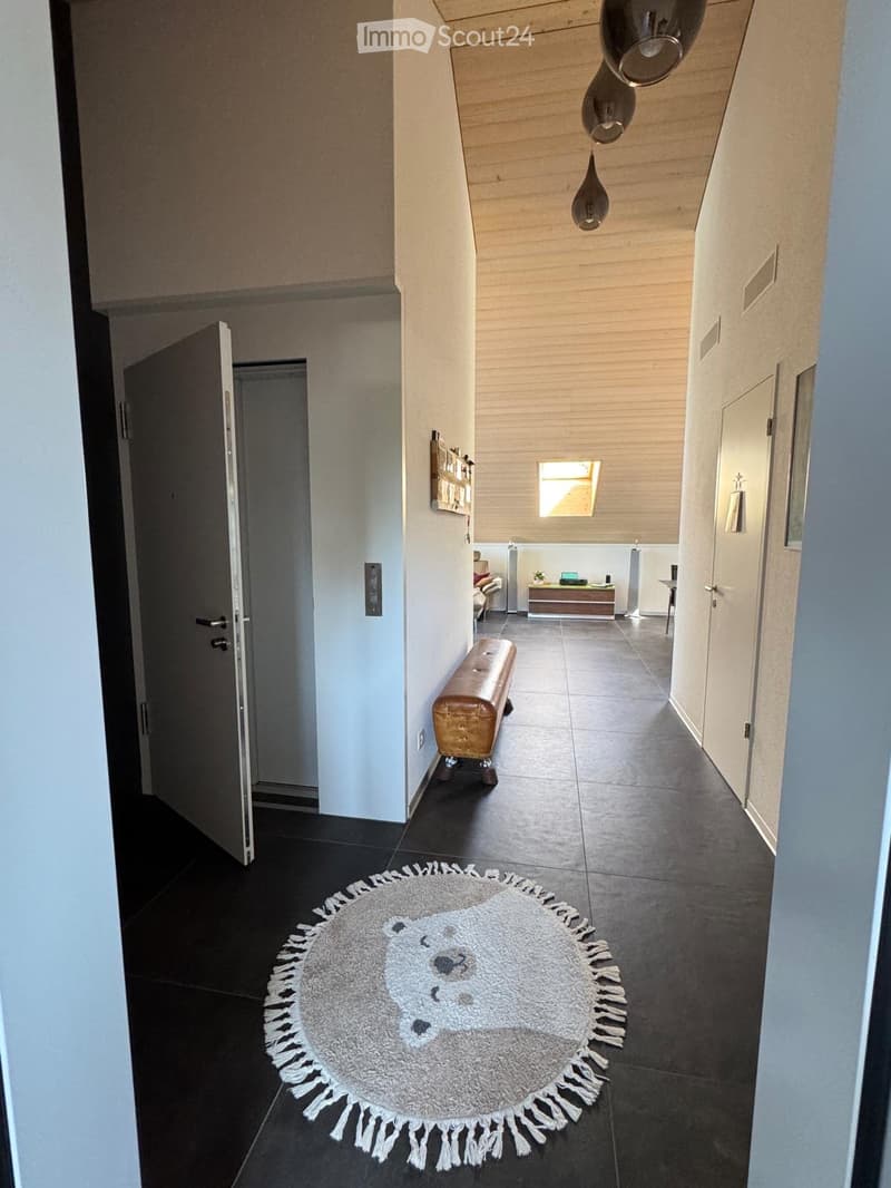 Modern hallway with dark tiled flooring, white walls, and a round rug with fringe detailing. Features a wooden bench, pendant lights, and an open doorway leading to another room.