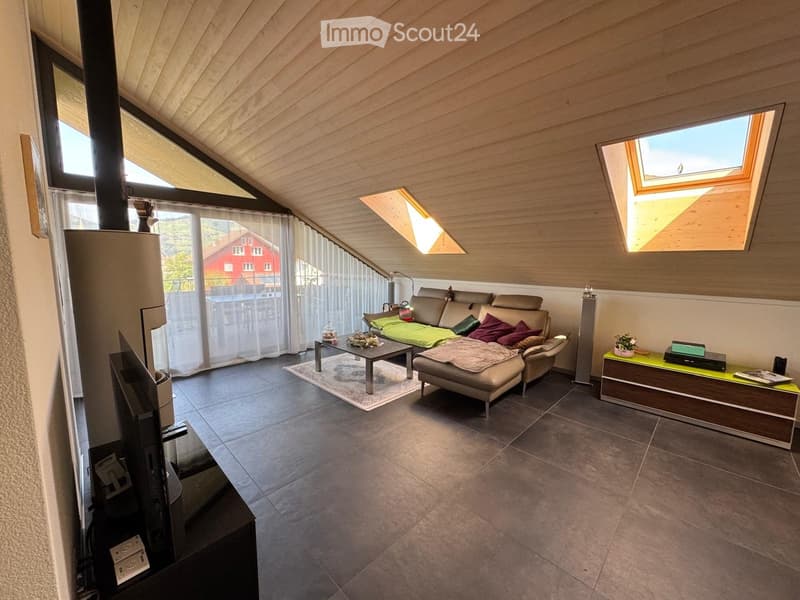 Modern living room with a slanted wooden ceiling, large windows, and skylights. Features a beige sectional sofa with colorful cushions, a coffee table, a side table with a potted plant, and a television on a stand.