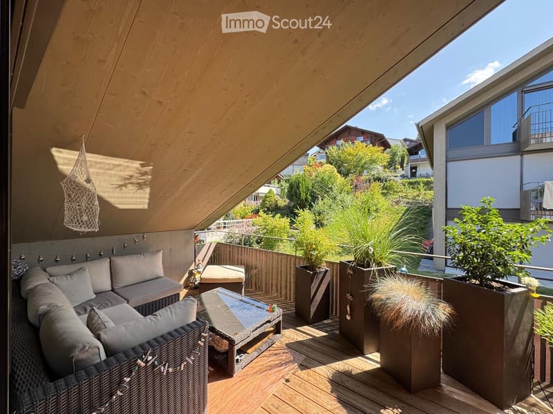 Balcony with wooden decking, modern furniture including a sofa and coffee table, and several large potted plants.