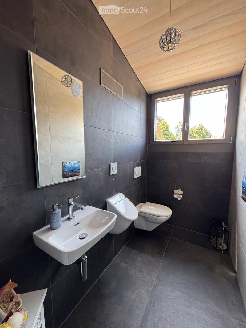 Modern bathroom with dark grey tiles, white sink, toilet, bidet, and a large mirror with a decorative picture. Features a window with a view of greenery.