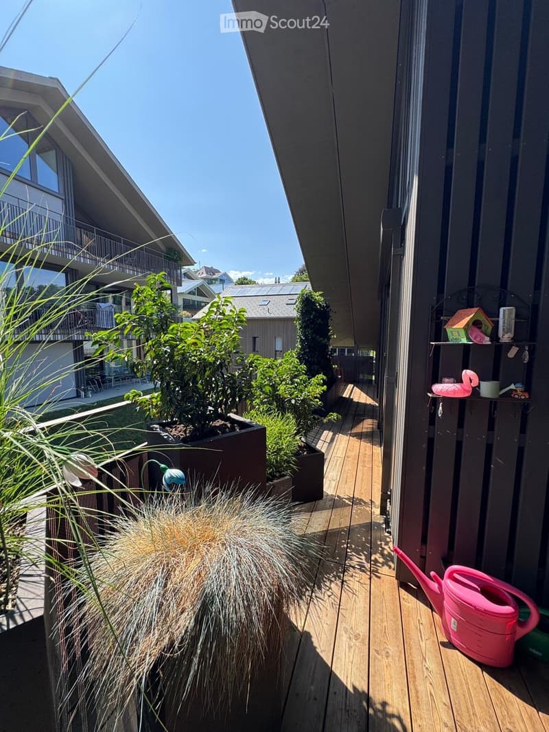 Balcony with wooden decking, potted plants, and a pink watering can. The view shows neighboring buildings and greenery.