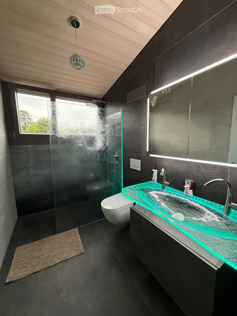 Modern bathroom with dark tiles, a glass shower enclosure, a floating sink with a green illuminated countertop, and a toilet. Features include a large mirror with built-in lighting and a pendant light.