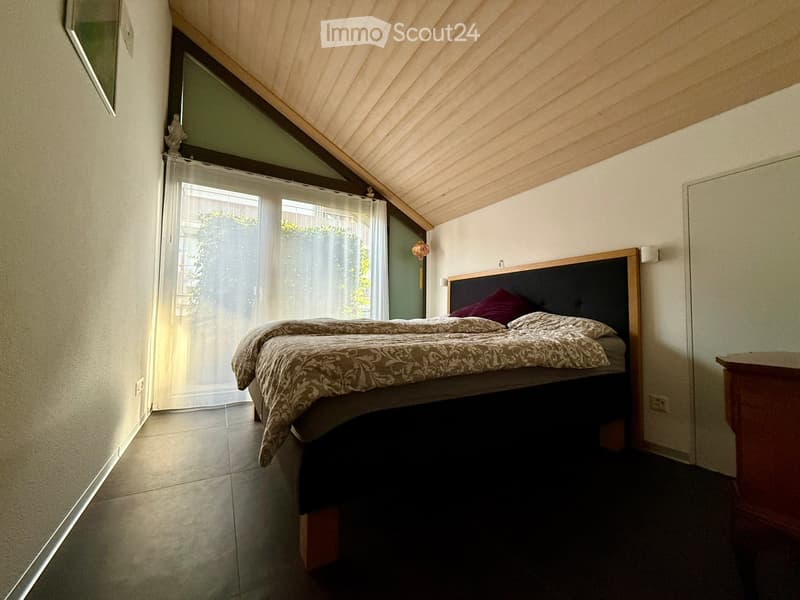 Cozy bedroom with a wooden bed, patterned bedspread, pillows, and a nightstand. Large window with sheer curtains allows natural light to enter.