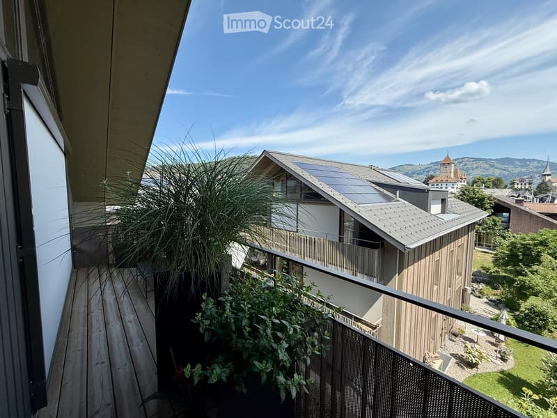 Balcony with wooden decking, metal railing, and lush green plants, overlooking a scenic town with historic buildings and solar panels on rooftops.