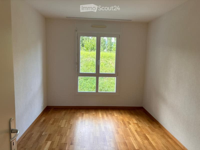 Empty room with wooden flooring and a large window showing a green outdoor view.
