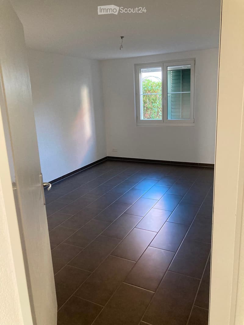Empty room with dark tiled floor and white walls, featuring a window with blinds and a door visible on the left side.