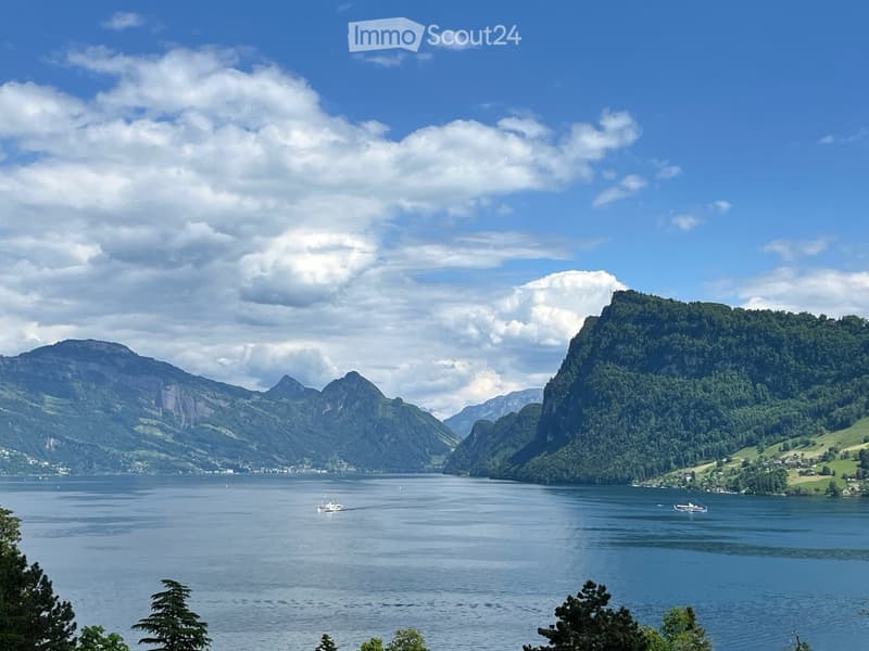 A scenic view of a lake surrounded by lush green mountains and a partly cloudy sky. Two boats are visible on the water, and the landscape includes dense forests and grassy slopes.