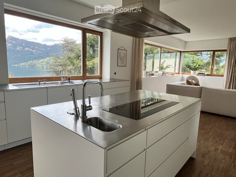Modern kitchen with white cabinets, stainless steel island, double sink, and large windows offering a scenic lake view. A small seating area with a sofa is visible in the background.