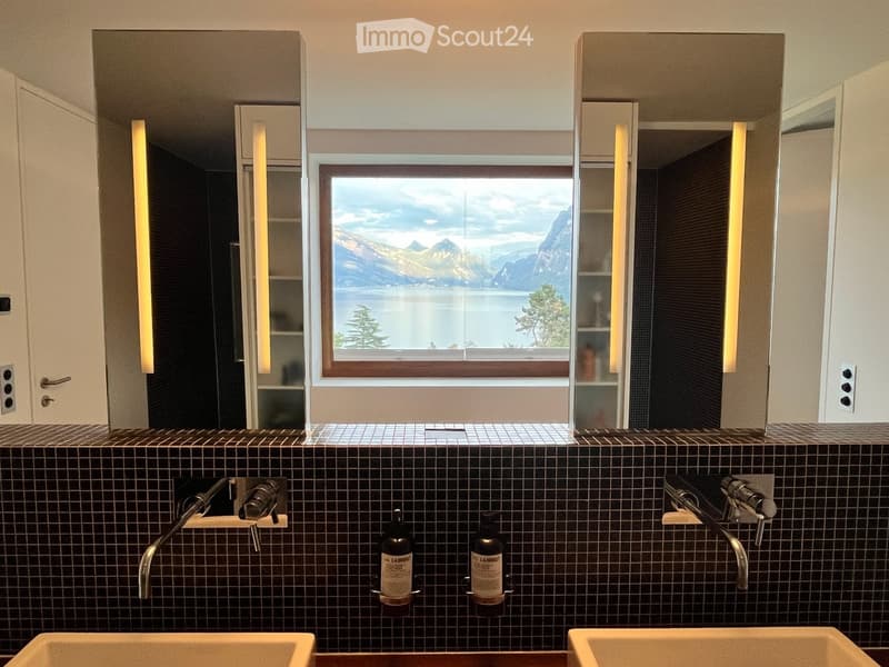 Modern bathroom with double sinks, large window overlooking a scenic lake and mountains, sleek mirrors with built-in lighting, and dark tiled walls.