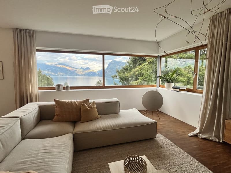 Modern living room with large windows offering a scenic mountain and lake view. Features a beige sectional sofa, coffee table, floor lamp, and decorative vases on the windowsill.