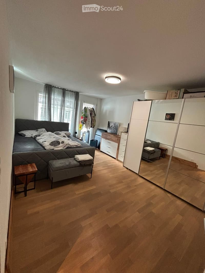 Modern bedroom with wooden flooring, large bed with dark headboard, nightstand, wardrobe with mirror, and bench at the foot of the bed.