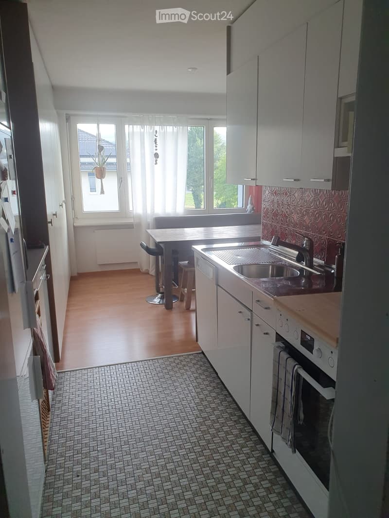 Modern kitchen with white cabinets, stainless steel sink, oven, and a small table with stools. The space features wooden flooring and large windows with sheer curtains.