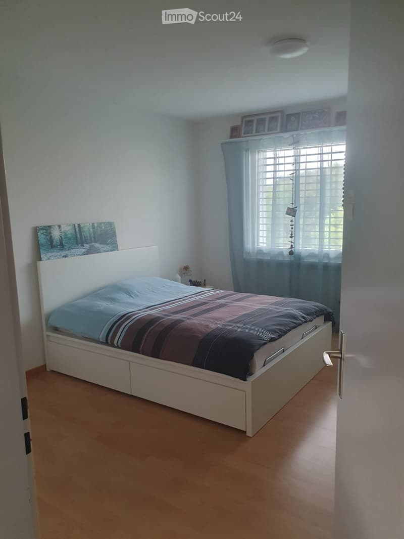 Bedroom with a white bed, striped bedding, a nightstand, and a large window with sheer curtains. The room has light wooden flooring and white walls.