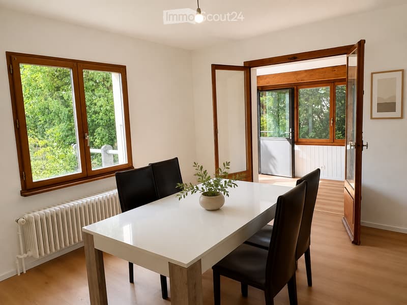 Bright dining area with a white table, four black chairs, and a small potted plant. Large windows with wooden frames offer a view of greenery, and the room has light wooden flooring and white walls.
