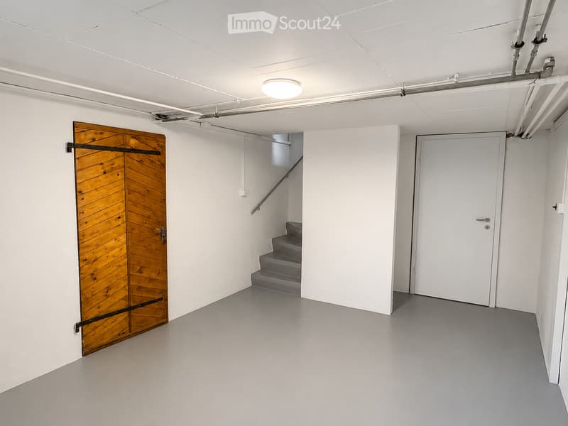 Empty room with white walls, grey floor, wooden door, white door, and a staircase leading upwards.