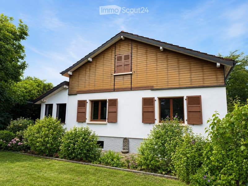 Two-story house with white walls, wooden accents, and brown shutters. The house features multiple windows and is surrounded by a well-maintained garden with green bushes and flowers.