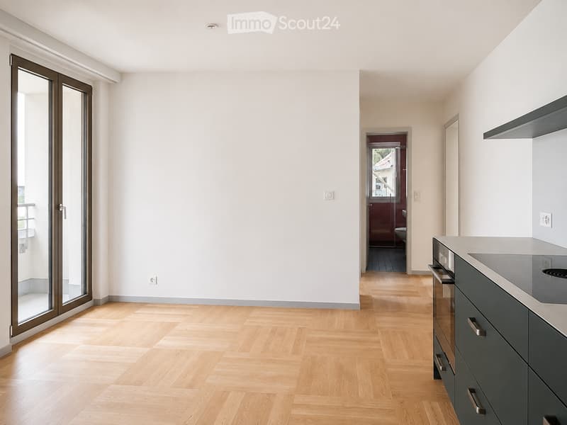 Modern, unfurnished room with light wooden flooring, white walls, and a sliding glass door leading to a balcony. A kitchen area with dark grey cabinets and a built-in oven is visible.