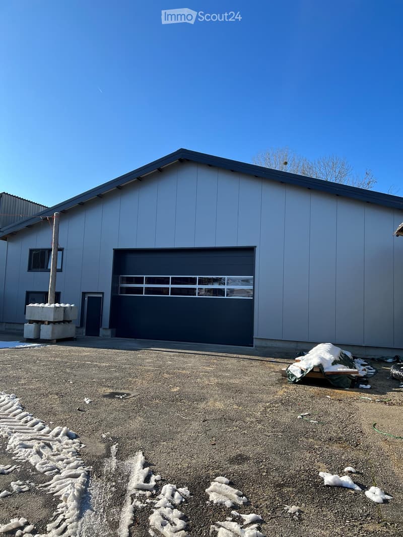 A modern, single-story building with a grey metal exterior, large garage door, and small windows. The surrounding area has gravel ground and scattered snow patches.