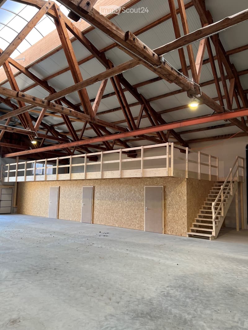 Large, open, unfinished space with exposed wooden beams, concrete floor, and a wooden staircase leading to an upper level with a wooden railing and several doors.