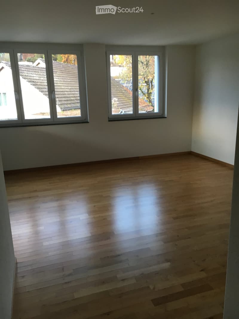 Empty room with light wooden flooring and large windows offering views of neighboring rooftops.