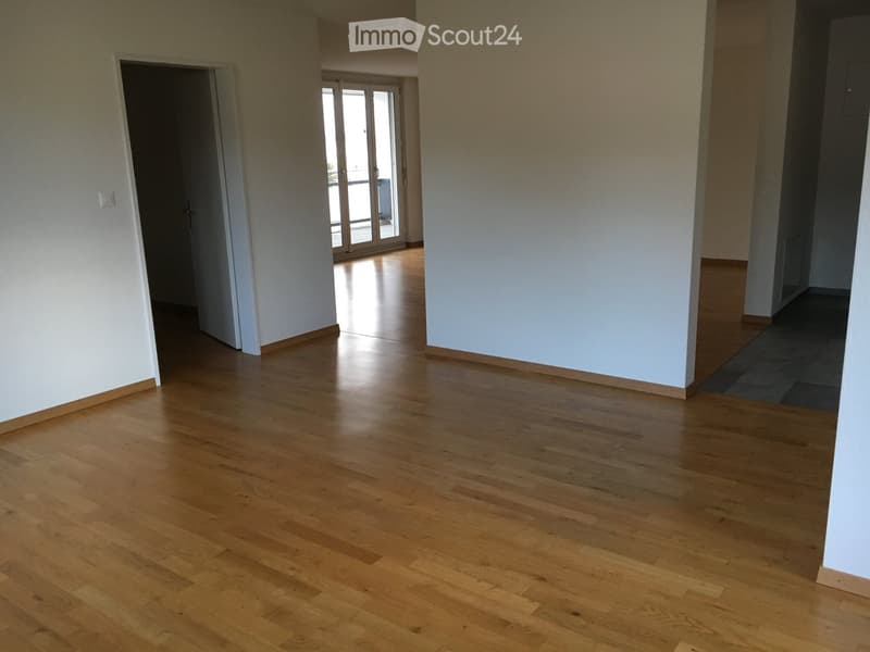 Empty room with light wooden flooring and white walls, featuring an open doorway leading to another room with large glass doors and a view of the outside.