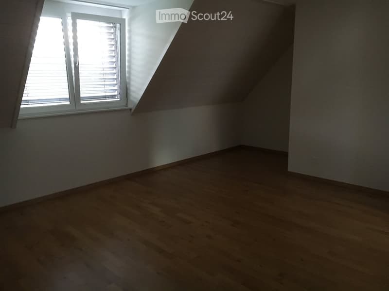 Empty attic room with wooden flooring, white walls, and a single window with blinds.
