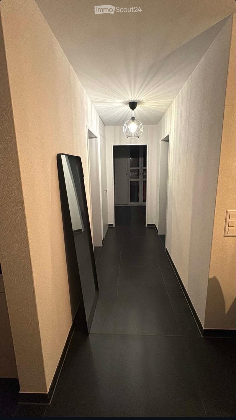 A long, narrow hallway with beige walls, dark tiled flooring, and a single hanging light fixture. The hallway leads to a door at the end.