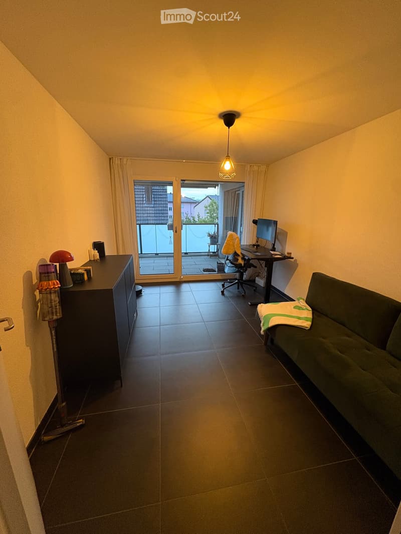 A modern living room with a dark green sofa, a desk with a computer, a hanging lamp, and a balcony with a view of the outside.