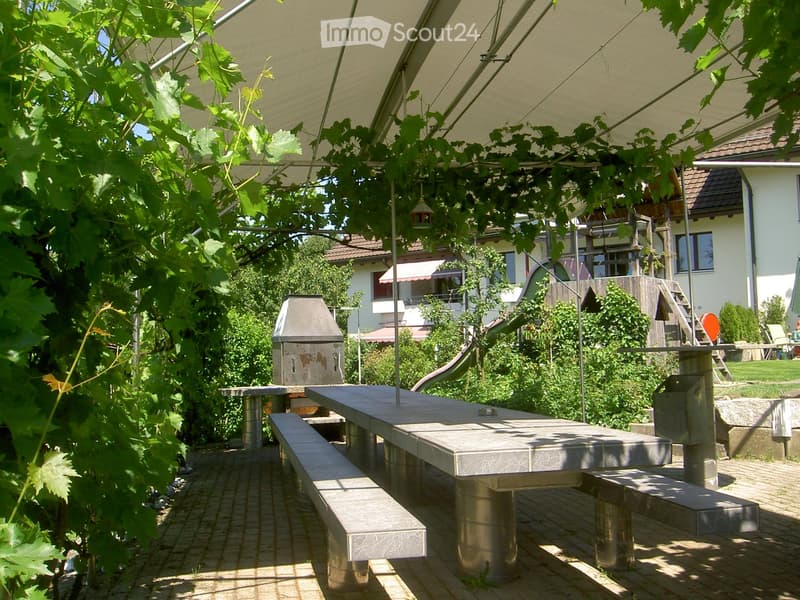 Outdoor dining area with a large table, benches, and a grill, surrounded by greenery and a covered canopy.