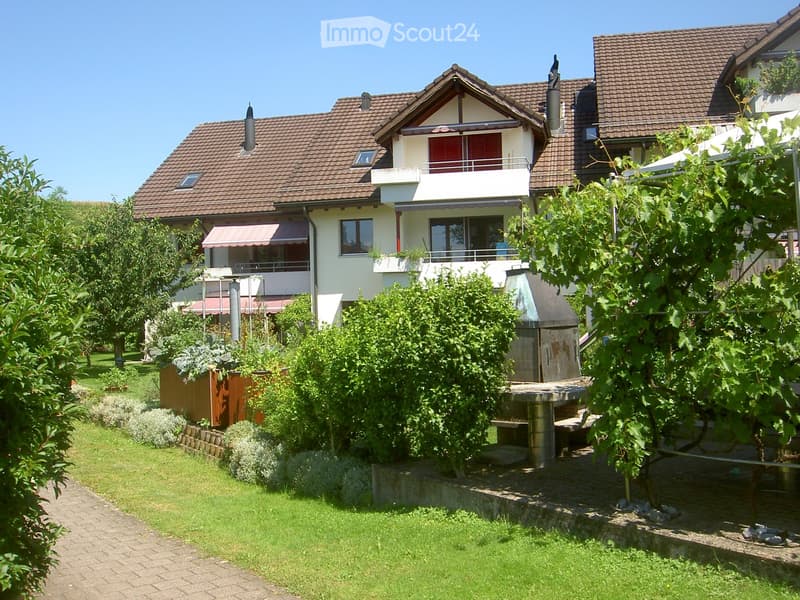 A well-maintained garden with a brick pathway, green lawn, various trees and shrubs, and a modern house with a balcony and red-tiled roof in the background.