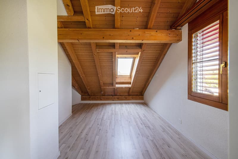 An empty attic room with wooden beams, wooden flooring, and large windows, including a skylight and a side window with blinds.