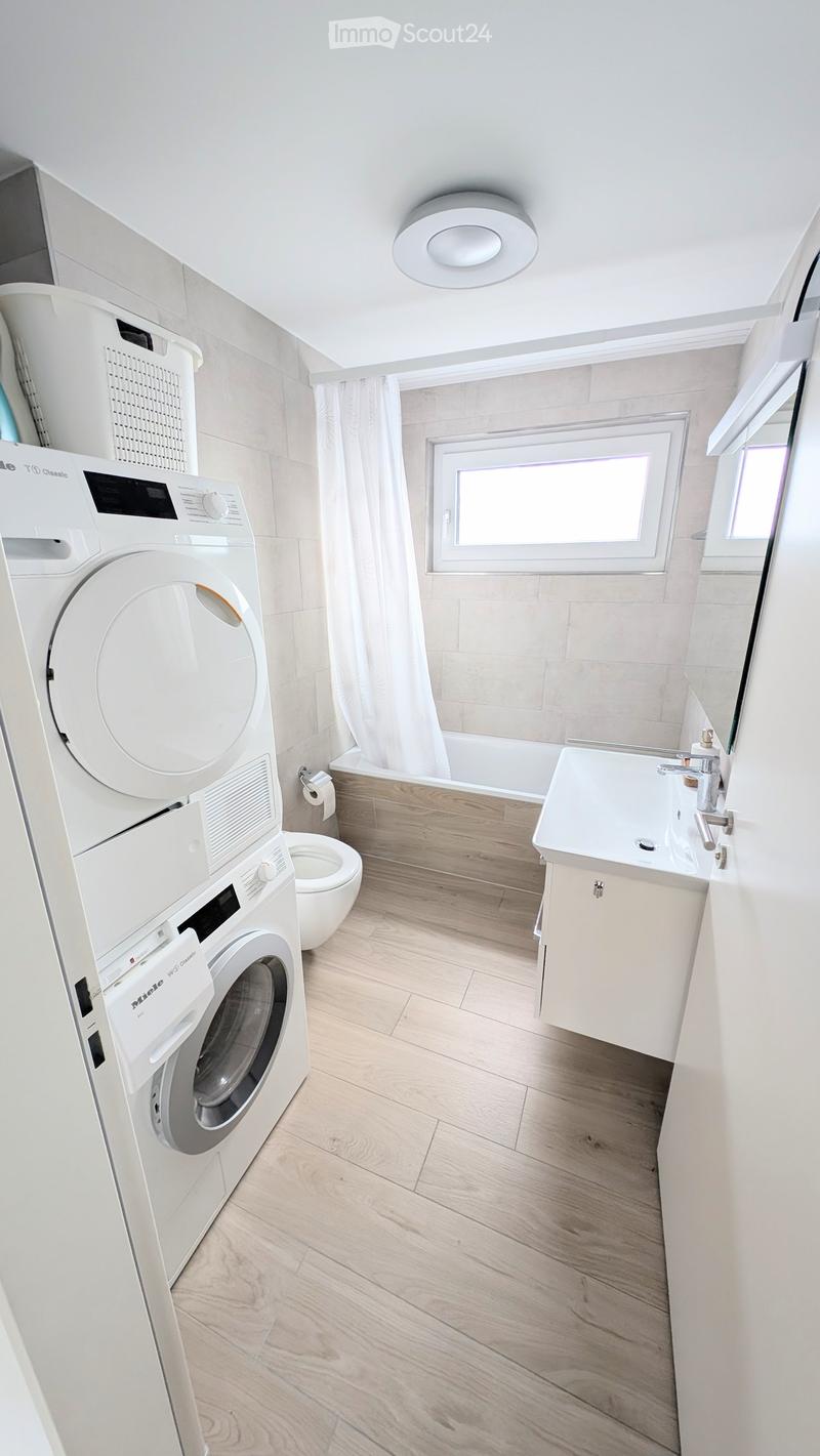 Modern bathroom with light wooden flooring, white tiled walls, a washing machine, toilet, bathtub, sink, and a window with a curtain.