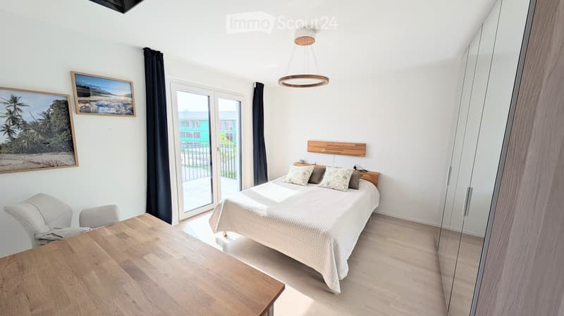 Modern bedroom with a double bed, nightstand, chair, small table, and large mirror. The room features light wooden flooring and white walls.
