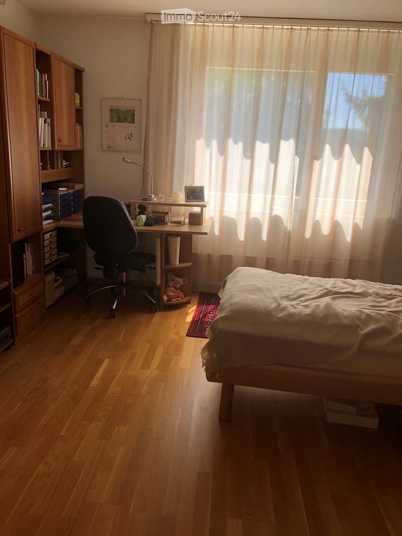 Bright bedroom with wooden flooring, a bed with a white blanket, a desk with a chair, a cabinet, and a window with sheer curtains.