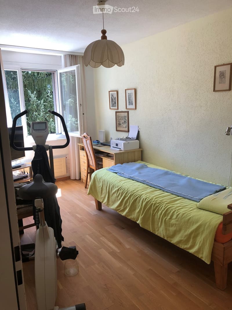 Well-lit bedroom with wooden flooring, a single bed with green and blue bedding, a desk with a chair, an exercise bike, and framed pictures on the wall.