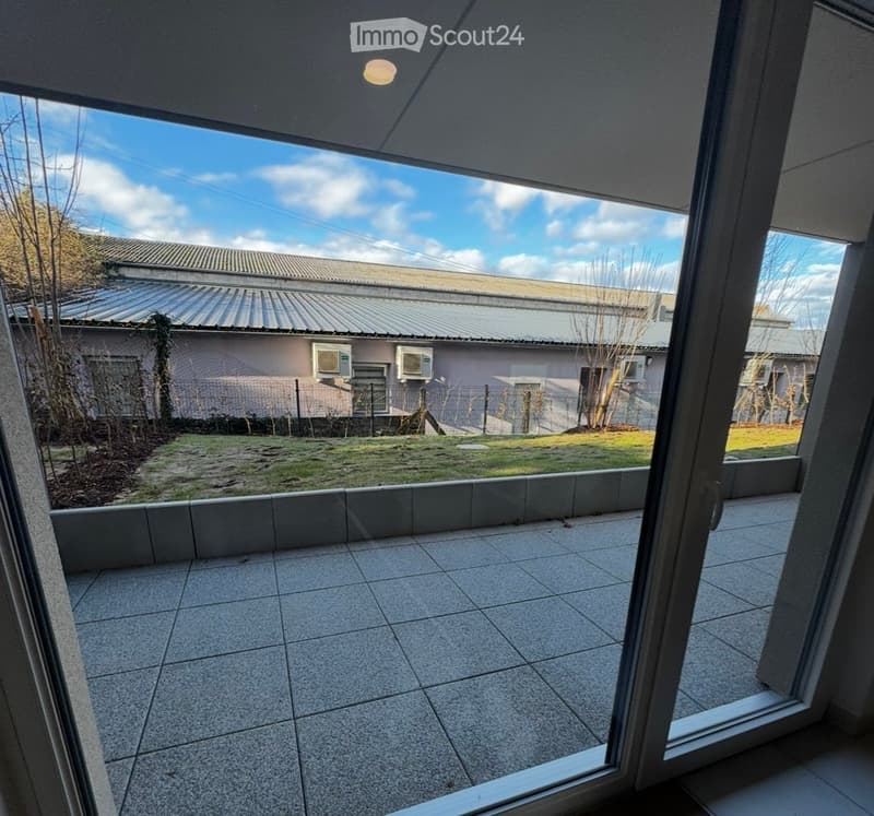 Balcony with tiled floor, open to a view of a grassy area and neighboring building with air conditioning units.