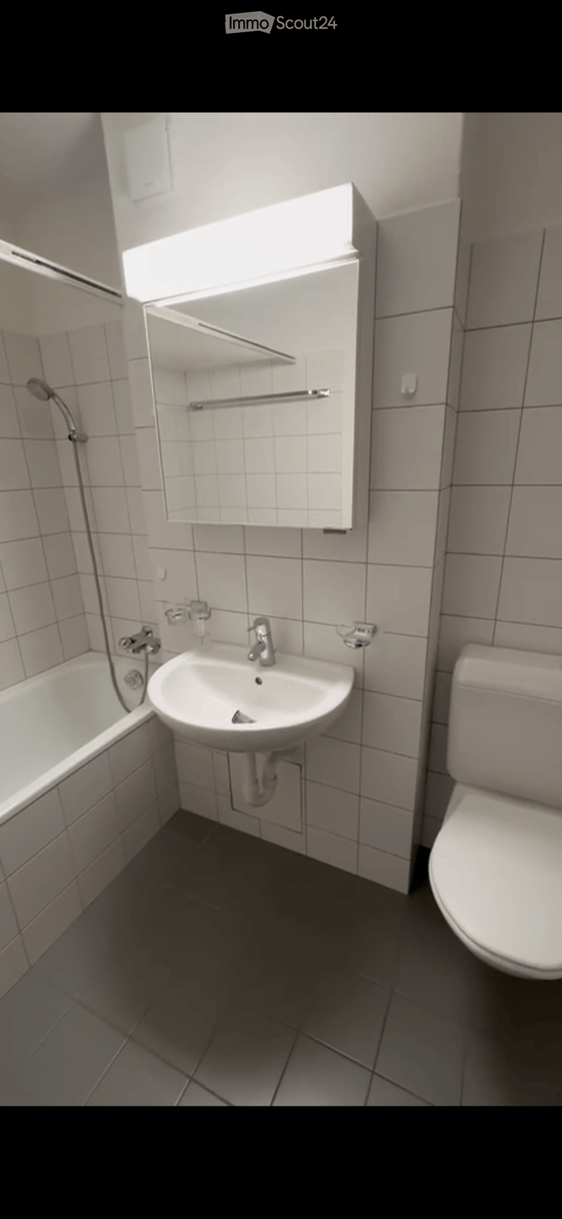 Modern bathroom with a bathtub, sink, toilet, mirror, and showerhead. The walls are tiled, and the floor is dark.