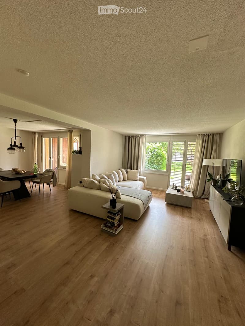 Modern living room with light wooden flooring, large windows with curtains, a white sectional sofa, a coffee table, dining area with chairs and table, and decorative items on side tables.