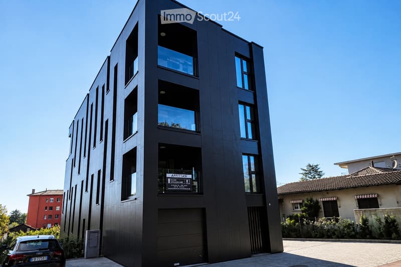 Modern, multi-story building with dark facade and large windows, featuring a garage entrance and a sign reading "AFFITTISI" (For Rent).