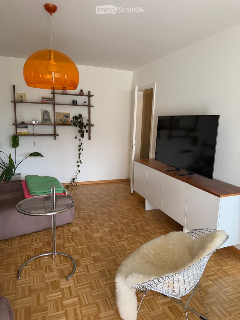 Modern living room with wooden herringbone flooring, a purple sofa, a round glass coffee table, a white wire chair with a fur throw, a large flat-screen TV on a wooden stand, and a wall-mounted shelf with books and decorative items.