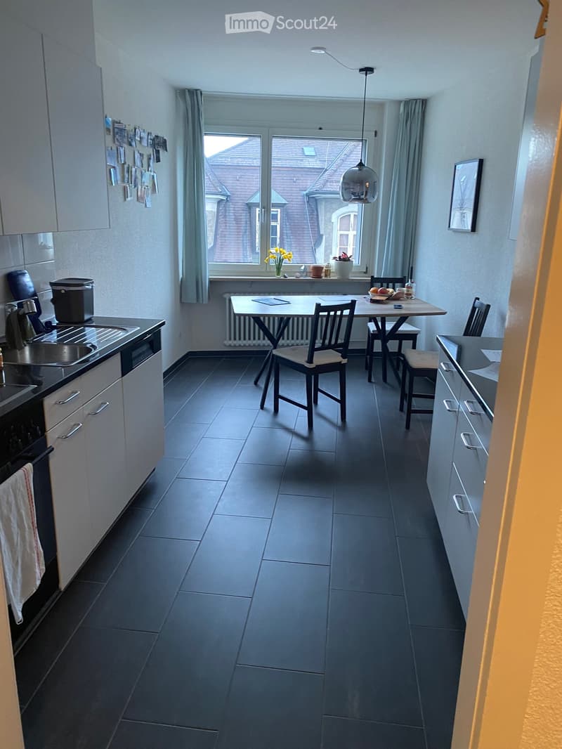 Modern kitchen with dark tiled floor, white cabinets, sink, oven, fridge, and a small dining area with a table and chairs. There is a window with a view of a brick house and some potted plants on the windowsill.