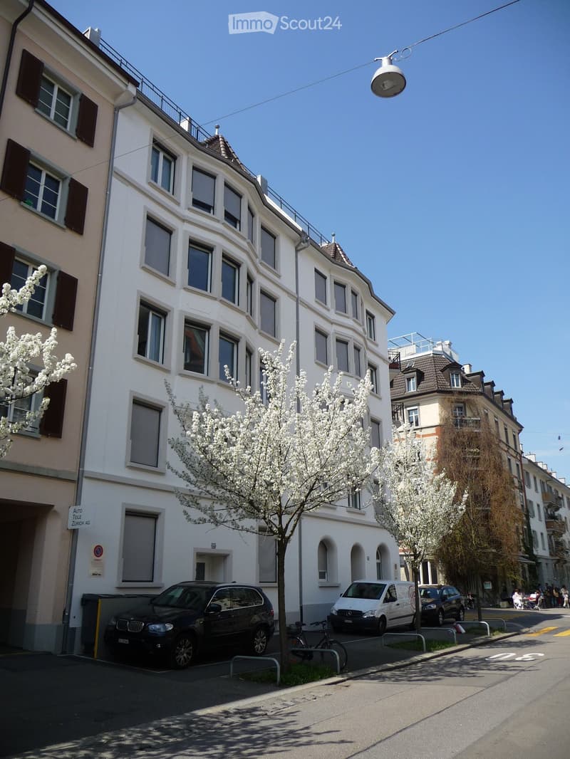 Multi-story white building with numerous windows and balconies, surrounded by blooming trees and parked cars.
