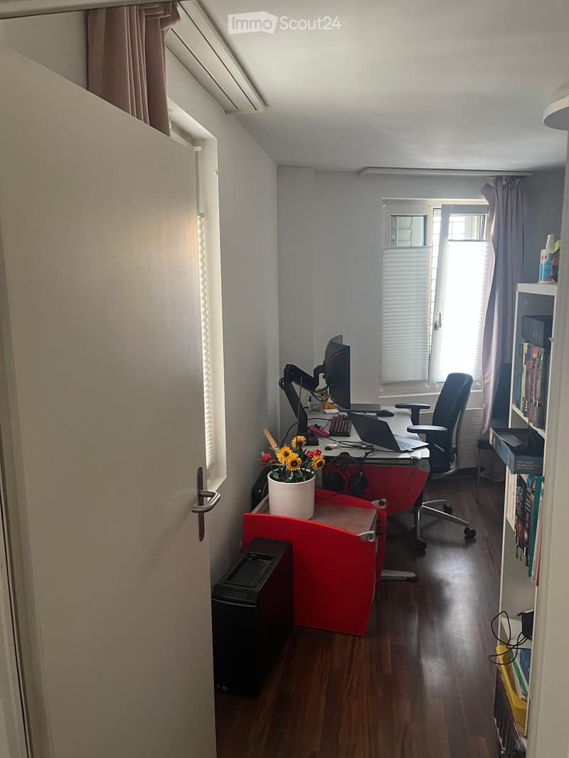 A modern, well-lit home office with a red desk, ergonomic chair, laptop, monitor, keyboard, mouse, flower pot, bookshelf, and trash can. The room features wooden flooring and large windows with blinds.