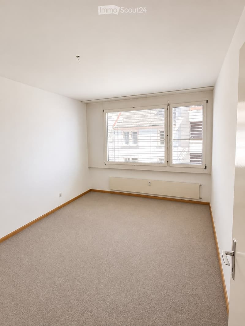 Empty room with white walls, carpeted floor, large window with blinds, radiator, and a door.